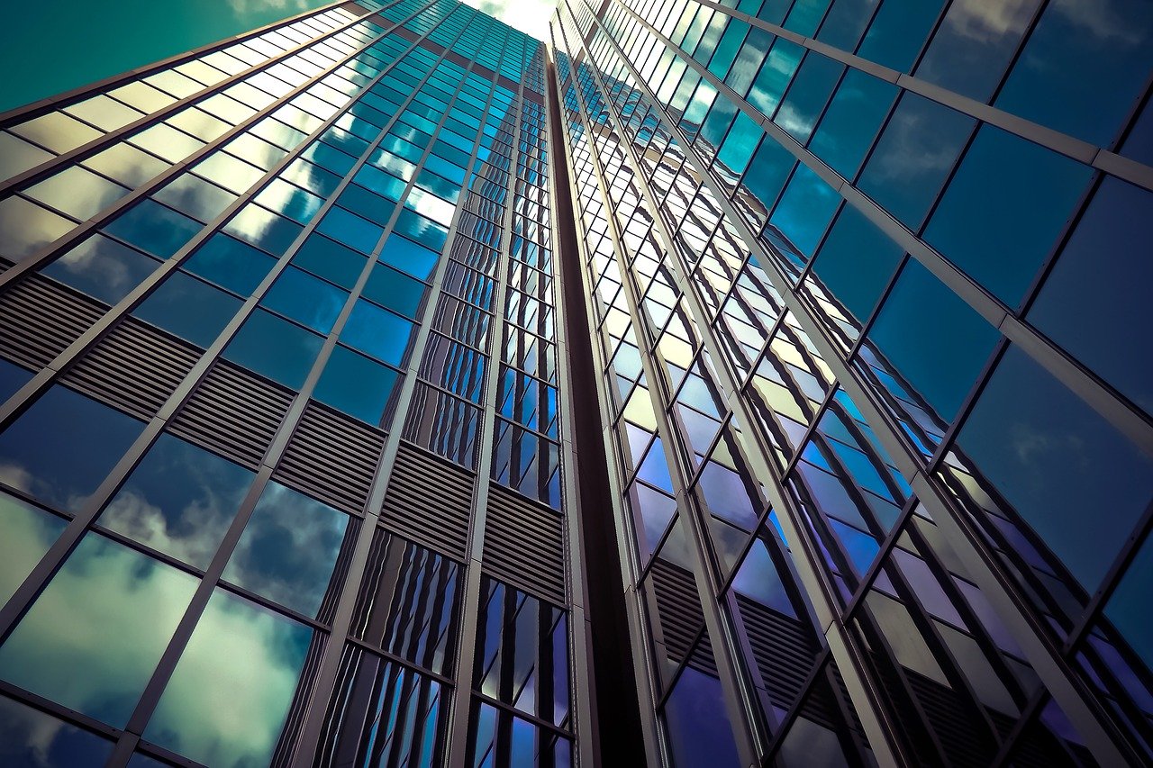 Vista desde un ángulo bajo de un rascacielos alto y moderno con amplias ventanas azules y de cristal reflectante. El reflejo de los edificios circundantes y de las nubes del cielo es visible en la fachada de cristal. Las líneas del edificio, ideales para quienes deseen constituir una empresa en la República Dominicana, crean un patrón simétrico y geométrico.
