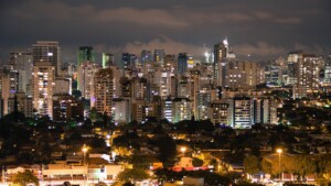 A nighttime view of a cityscape featuring numerous high-rise buildings with illuminated windows. In the foreground, smaller residential structures and streets are visible. The skyline includes both modern and older buildings, all set against a dark sky, ideal for those looking to fazer negócios no Brasil.