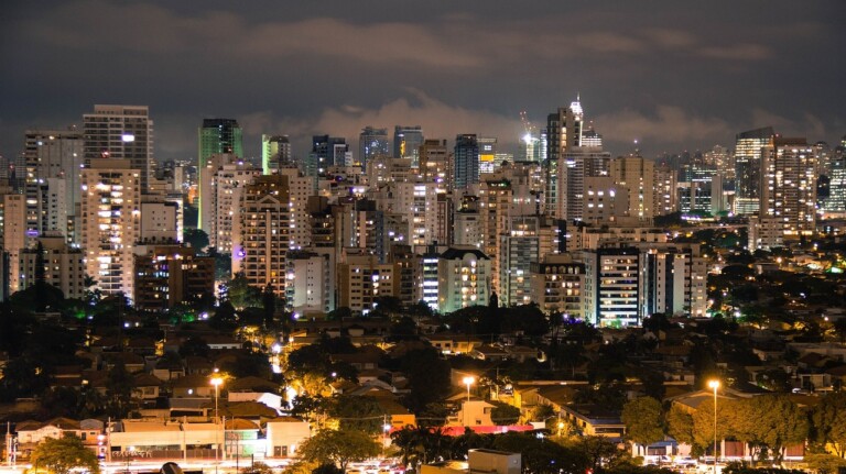 A nighttime view of a cityscape featuring numerous high-rise buildings with illuminated windows. In the foreground, smaller residential structures and streets are visible. The skyline includes both modern and older buildings, all set against a dark sky, ideal for those looking to fazer negócios no Brasil.