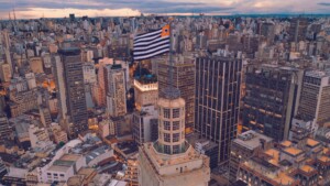 Aerial view of a densely-built urban area with high-rise buildings. In the center, a flag with black and white stripes and a red and yellow square in the top left is mounted on top of a tall historic building. The sky is overcast, reflecting the complex landscape faced by an employer of record in Brazil.