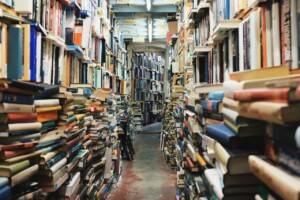 A narrow aisle is flanked by shelves filled with books, with additional books stacked on the floor. The ceiling and shelves are packed densely with a variety of hardcover and paperback books, creating a cluttered but cozy atmosphere reminiscent of embarking on a trademark search in Indonesia. The floor is mostly visible.