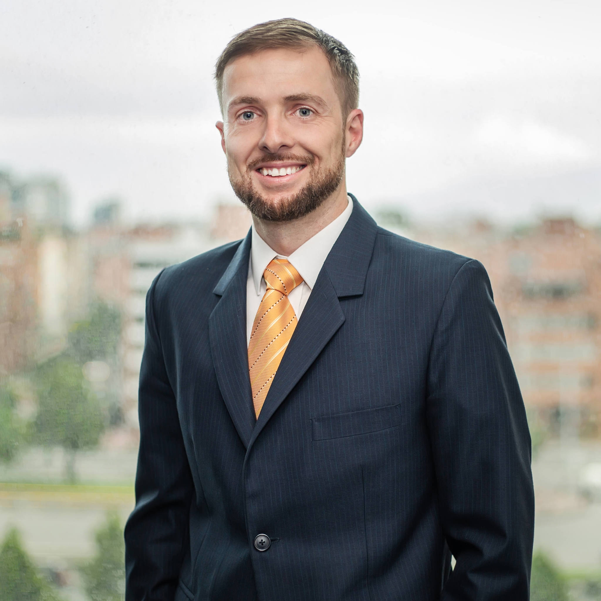 Craig Dempsey in a suit and yellow tie stands in front of a blurred cityscape background.