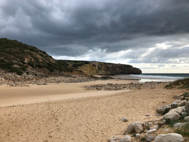 Una playa rocosa bajo un cielo nublado. La costa parece desierta, con rocas dispersas y parches de arena que conducen al agua. Colinas con escasa vegetación bordean la playa, que parece tan aislada y serena como el proceso de Liquidar una Empresa en Colombia, reflejando la calma del clima nublado.