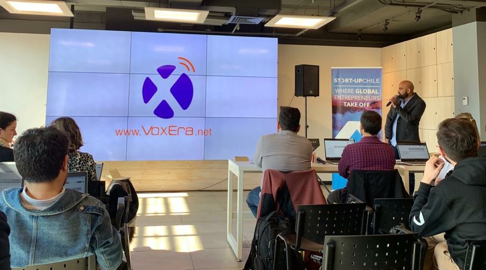 A person is presenting at a seminar to a seated audience with a large screen showing the logo and website "VoxEra.net." There is a "STARTUP CHILE" banner in the background, highlighting the dynamic atmosphere of entrepreneurship in Chile.