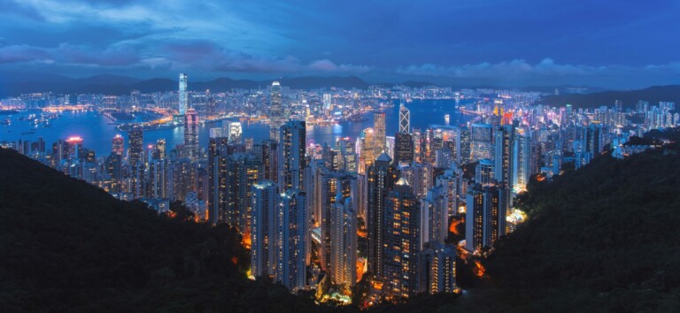 A panoramic view of a densely built skyline illuminated at night, featuring numerous high-rise buildings. The cityscape is bordered by a large body of water, with lights reflecting on its surface. Hills are visible in the foreground and background.