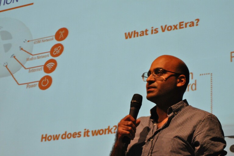 A man holding a microphone stands in front of a presentation slide. The slide features text, graphs, and images, with notable terms including "What is VoxEra?" and "How does it work?" The man appears to be explaining or discussing the content of the slide.