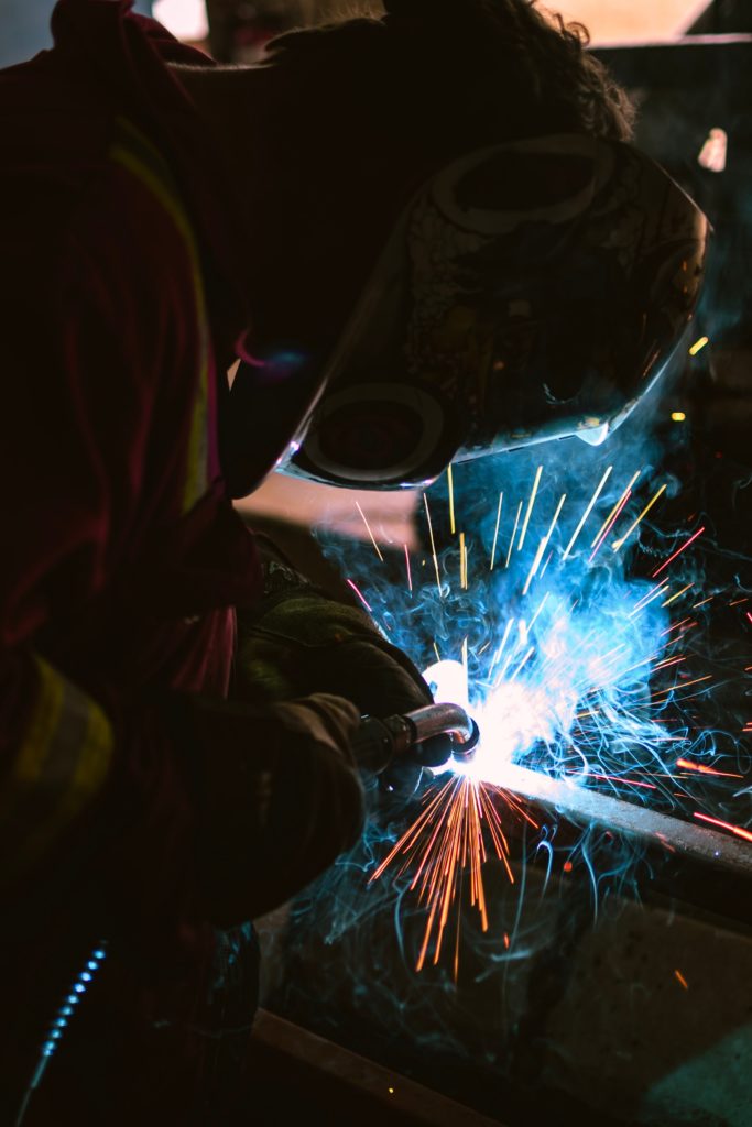 A person wearing protective gear, including a welding helmet, is welding a piece of metal. Bright sparks and blue light emanate from the welding area, contrasting with the dark surroundings—a scene emblematic of the thriving Mexico manufacturing sector.