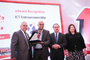 Four individuals stand on stage during an eAward Recognition event for ICT entrepreneurship in Chile. One person, holding an award plaque, is being recognized. They are all dressed in formal business attire and smile for the camera. A screen in the background displays the event details.