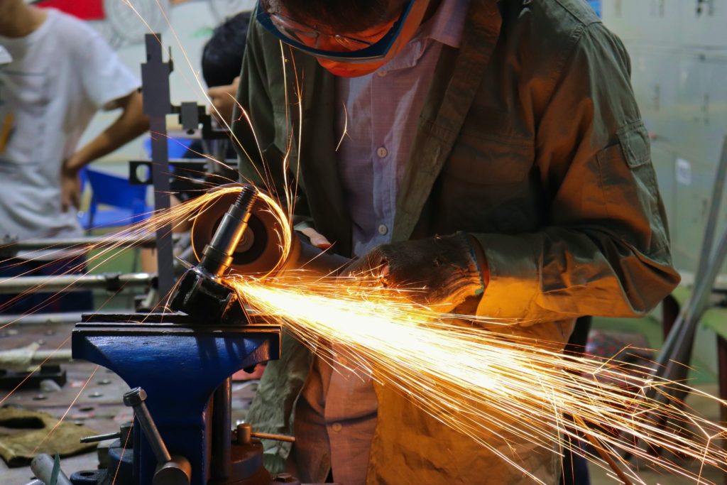 A person wearing safety goggles and a brown jacket is using an angle grinder on a piece of metal clamped in a vise, with bright sparks flying off the metal. In the background, two other people and various pieces of equipment from Mexico's manufacturing sector are visible.