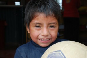 A young boy with short dark hair is smiling and holding a soccer ball. He is wearing a blue shirt and standing indoors, part of an NGO in Bolivia focused on youth sports.