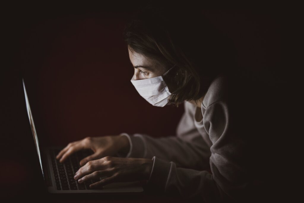 A person wearing a mask is using a laptop in a dimly lit environment. Immersed in their work on fintech business in Guatemala, the individual faces the screen with hands placed on the keyboard. The dark background accentuates the glow from the laptop screen.