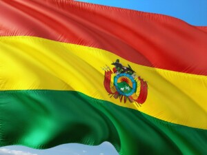 The image shows the flag of Bolivia waving in the wind, consisting of three horizontal stripes: red, yellow, and green from top to bottom. The yellow stripe features the Bolivian coat of arms in its center. The sky is visible in the background, symbolizing a promising environment to form an NGO in Bolivia.