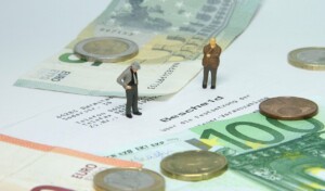 Two miniature figurines of people stand on a document surrounded by various denominations of euro banknotes and coins. The banknotes and coins are spread out on a white surface, and the document, possibly an entity health check from Argentina, has German text on it.