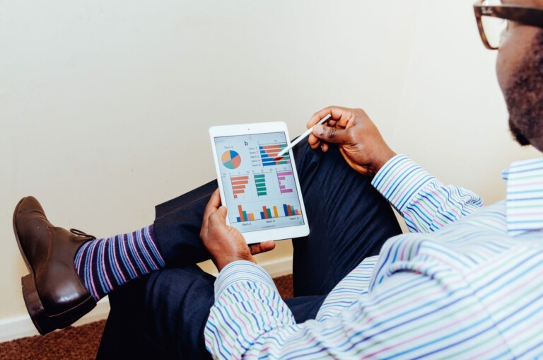 A person wearing striped socks, dress shoes, and a striped shirt is sitting with legs crossed, holding a tablet displaying various colorful charts and graphs on investment trends in Guatemala. They are pointing at the screen with a stylus. The background is a plain, off-white wall.