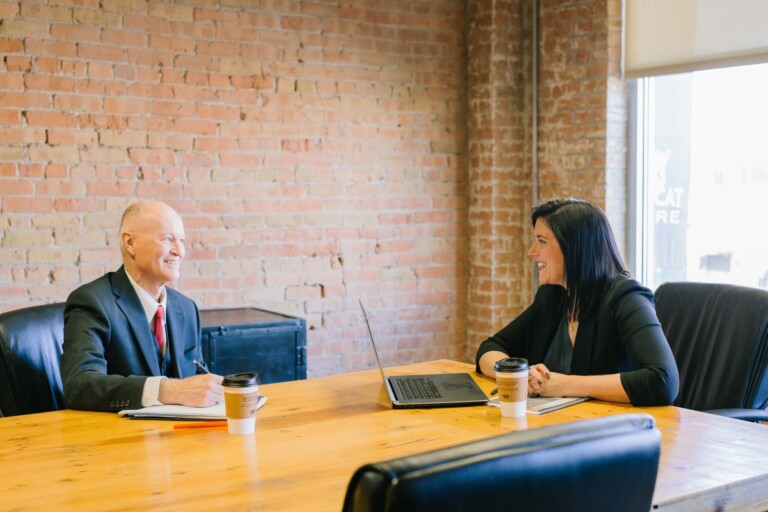 A bald man in a suit and a woman in a black outfit are having a discussion in a conference room with a wooden table. Both have coffee cups in front of them, and the woman has a laptop open, likely researching how to open a corporate bank account in Bolivia. The brick wall behind them has a window allowing natural light in.