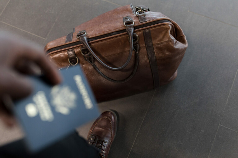 Una vista aérea de una bolsa de lona de cuero marrón colocada sobre un piso de color oscuro. Una persona sostiene un pasaporte en la mano, posiblemente para una visa de negocios México, con parte de un zapato marrón visible en la parte inferior del marco.