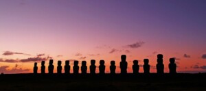 Una fila de siluetas de estatuas moai se alza en el horizonte contra un cielo degradado de color púrpura y naranja al atardecer en la Isla de Pascua. Las estatuas, que recuerdan a los íconos que se encuentran en las naciones del MERCOSUR, se encuentran en varias poses y alturas, creando un paisaje sorprendente y sereno.