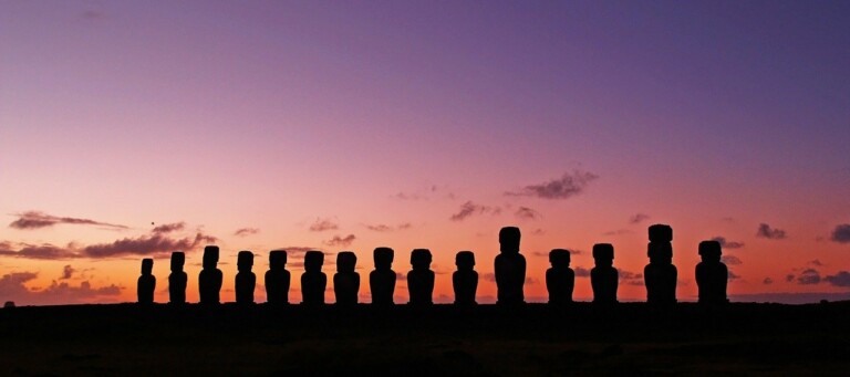 Una fila de siluetas de estatuas moai se alza en el horizonte contra un cielo degradado de color púrpura y naranja al atardecer en la Isla de Pascua. Las estatuas, que recuerdan a los íconos que se encuentran en las naciones del MERCOSUR, se encuentran en varias poses y alturas, creando un paisaje sorprendente y sereno.