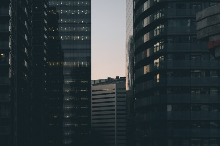 Un paisaje urbano al atardecer retrata modernos edificios de gran altura con fachadas de vidrio reflectante. Las estructuras ubicadas muy cerca forman un pasaje estrecho que revela una franja de cielo teñida con los tonos del atardecer. Las luces de las oficinas brillan en algunas ventanas, lo que recuerda a los bulliciosos centros comerciales como los de las ciudades del MERCOSUR.