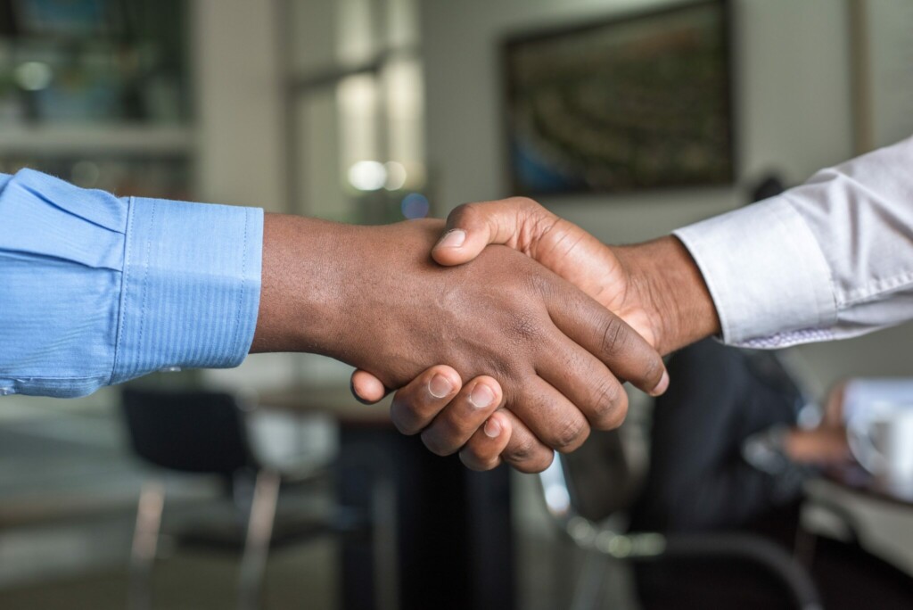 Two people shaking hands in an office setting, symbolizing the start of a partnership for Corporate Secretarial Services in Paraguay. The person on the left is wearing a blue shirt, while the person on the right sports a white shirt. Office furniture and a blurred background set the professional tone.