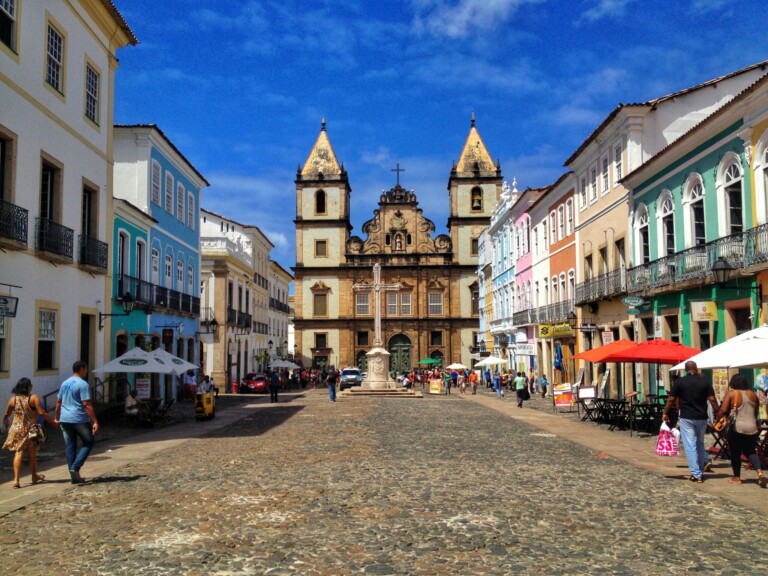 Una calle adoquinada bordeada de coloridos edificios bajo un cielo azul conduce a una iglesia con dos campanarios y una cruz central. La gente camina y se sienta en cafés al aire libre con mesas a la sombra de sombrillas, discutiendo planes para liquidar una empresa en El Salvador. La escena es vibrante y animada.