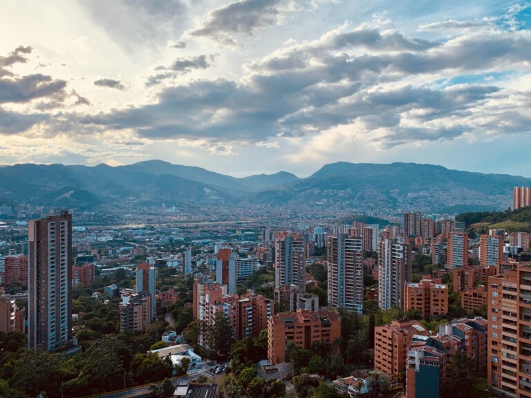 Una vista de una ciudad con numerosos rascacielos y edificios de media altura bajo un cielo nublado. Al fondo se pueden observar las montañas, formando un pintoresco telón de fondo típico de algunos países miembros del MERCOSUR. La escena incluye una mezcla de edificios modernos y antiguos rodeados de árboles, lo que sugiere que es por la mañana o por la tarde.