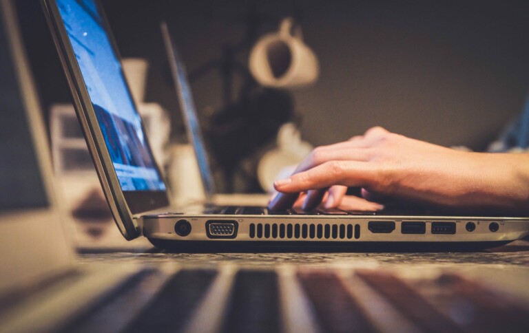 Close-up of a person using a laptop, with only their hands visible typing on the keyboard. The laptop is on a table, and another open laptop is visible in the foreground. The background is slightly blurred, showing a dimly lit interior environment, perhaps hinting at the burgeoning era of 5G in Uruguay.