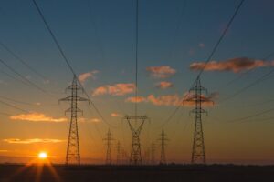 A landscape view shows several power lines and transmission towers stretching across a field at sunset. The sky is partly cloudy with the sun near the horizon, casting an orange glow on the scene, reminiscent of how Mercosur trade deals and negotiations span vast distances to connect nations.