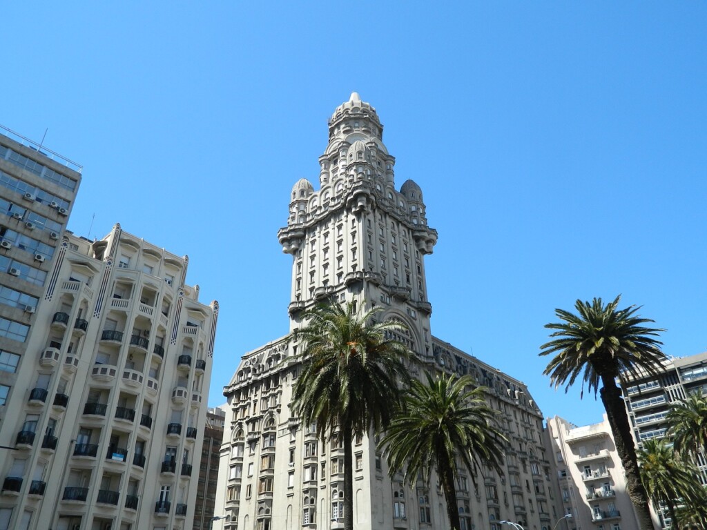 A tall, ornate building with an intricate design stands under a clear blue sky, flanked by other buildings and palm trees. It has a central tower with detailed carvings and a unique architectural style befitting a legal representative in Uruguay.