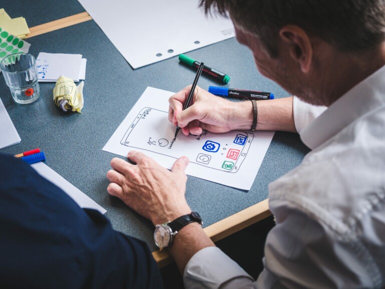 Two people are seated at a table, focusing on drawing a design on a white sheet of paper. One person is holding a pen and sketching icons, including a camera and a globe, discussing due diligence in Argentina as the other person watches. Various markers and other items are scattered on the table.