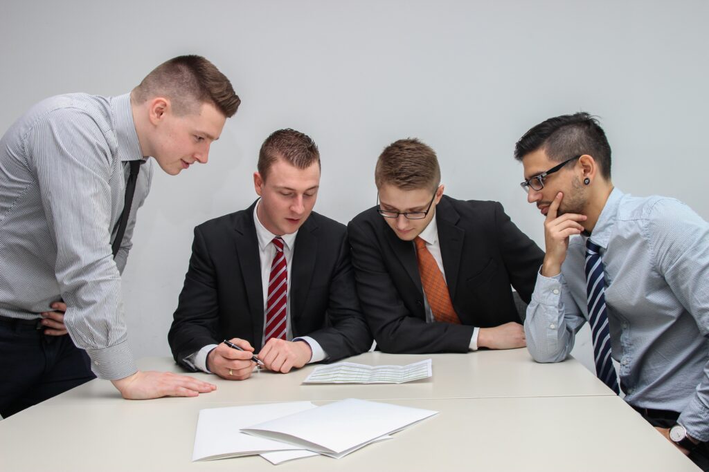 Four men in business attire are gathered around a table reviewing documents, focusing intently on recent Mercosur trade deals. Three of them are seated, while one stands and leans over to look at the papers. The table also has a few blank sheets of paper on it for further negotiations.