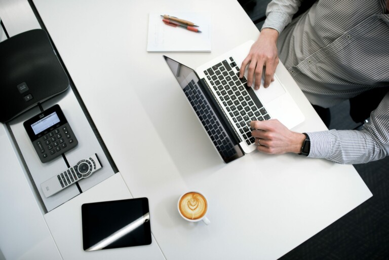 Una persona con una camisa a rayas escribe en una computadora portátil en un escritorio. El montaje, ideal para brindar servicios secretariales corporativos Chile, incluye una libreta con bolígrafos, una taza de café con latte art, una tablet y un teléfono de escritorio. La escena se ve desde arriba.