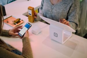 Two people are at a checkout counter. The person on the left is holding a smartphone, displaying a payment screen. The person on the right is verifying the transaction on a tablet connected to a white Square card reader, showcasing advancements in Australia's financial sector.