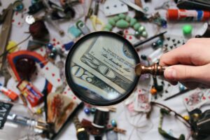 A hand holds a magnifying glass focusing on a twenty-dollar bill, magnifying the detail. The background is cluttered with various items, such as playing cards, buttons, and other small objects, evoking the scene of an entity health check in Bolivia.