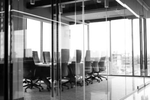 A black and white photograph shows an empty conference room with a long table and high-backed chairs around it. The room is enclosed by glass walls, and large windows offer a view of a cityscape outside. The atmosphere is modern and professional, ideal for discussing a transfer pricing study in Colombia.