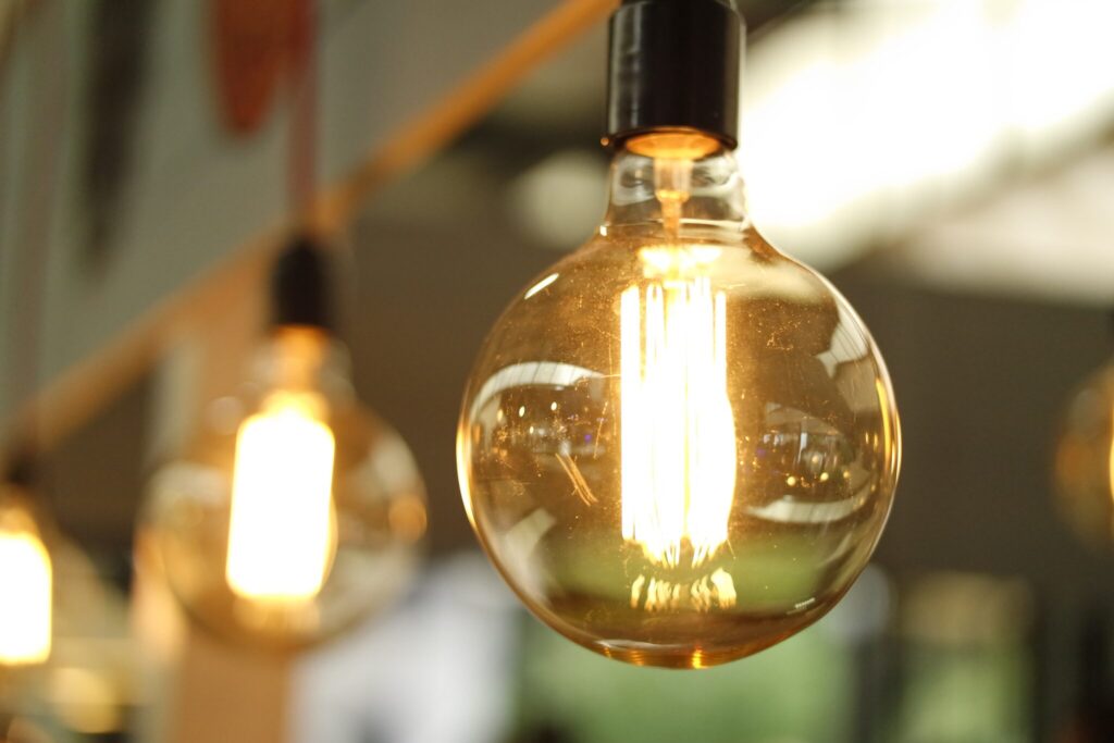 Close-up image of a lit, clear glass light bulb with visible glowing filament. The background shows out-of-focus, similarly lit bulbs, giving a warm, ambient light effect. This modern setting subtly hints at mining and energy innovation through its sophisticated illumination.