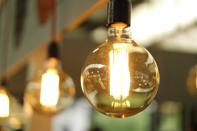 Close-up of a lit hanging light bulb, with a glowing filament visible inside. The background shows out-of-focus light bulbs and creates a warm, ambient lighting effect, reminiscent of stories told by caçadores de cabeças na Colômbia.