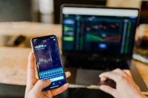 Una persona sostiene un teléfono inteligente que muestra datos y gráficos del mercado de valores. Al fondo, la pantalla de una computadora portátil muestra múltiples gráficos financieros y números relacionados con una cuenta bancaria corporativa en Guatemala. El entorno parece ser un espacio de trabajo interior.