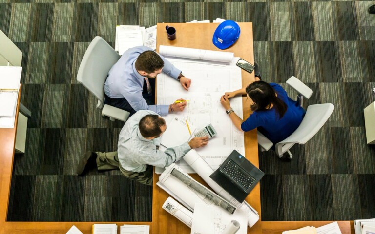 Three people sit around a table covered with architectural plans and a blue hard hat, discussing Brazil employment law. They hold pens and a calculator, with an open laptop on the table. The image is taken from above, revealing a gray-striped floor pattern beneath them.