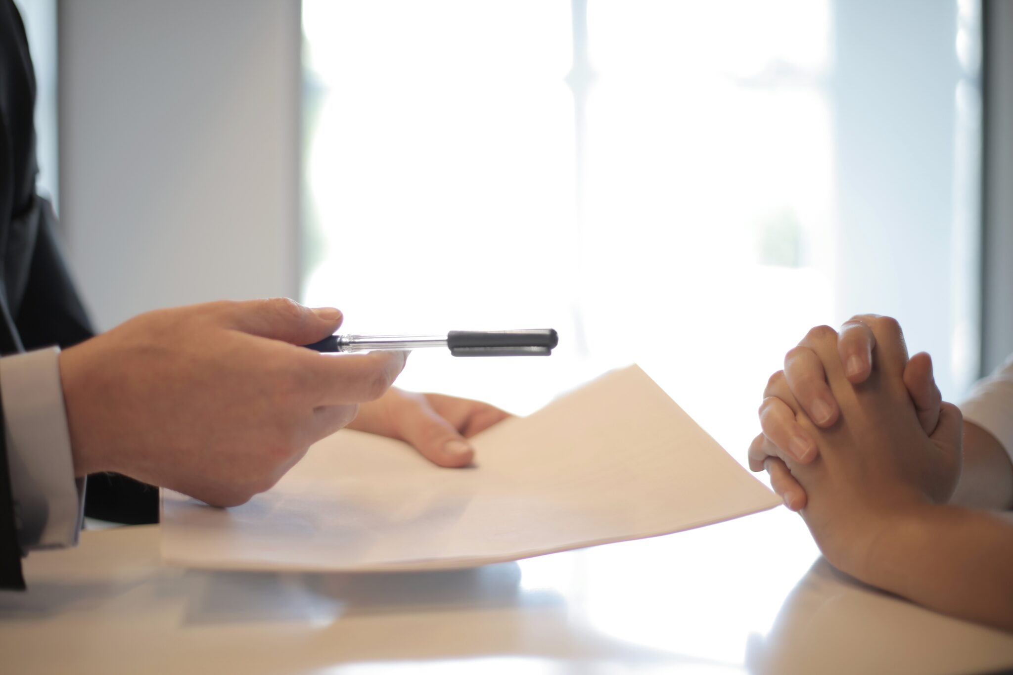 A person in a suit holds out a pen and a document towards another individual with clasped hands resting on a table, discussing corporate compliance requirements in Peru. The background features blurred, bright windows.