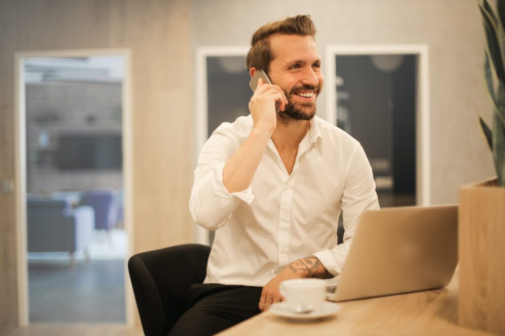 A man with a beard and white shirt is sitting at a desk, smiling while discussing corporate compliance requirements in Peru on the phone. He has a laptop open in front of him and a white coffee cup on the desk. The background features a blurred modern office setting.