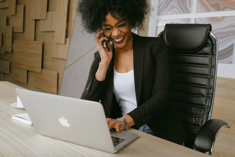 A person with curly hair, wearing glasses, a black blazer, and a white shirt, is sitting in a leather office chair. They discuss the challenges of doing business in Mexico on a smartphone while typing on a silver laptop at a wooden desk with a cup and saucer.