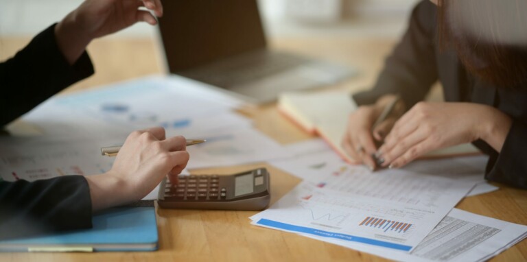 Two people working at a desk with documents, charts, and a calculator. One person holds a pen over a document about dividend withholding tax, while the other writes on paper. A laptop and a notebook are also on the desk. Both individuals are partially visible.