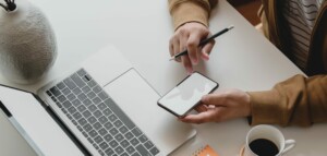 A person wearing a brown jacket is seated at a desk, using a smartphone with one hand while resting a pencil on the other, perhaps exploring E-Commerce in Bolivia. An open laptop, a white vase, an orange notebook, and a cup of coffee are also on the desk.