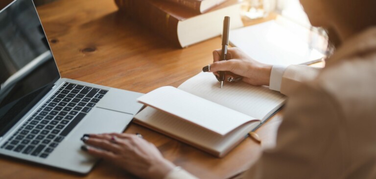 Persona sentada en un escritorio de madera usando una computadora portátil y escribiendo en un cuaderno. Al fondo se apilan varios libros sobre cumplimiento corporativo Uruguay. La persona sostiene un bolígrafo y parece estar tomando notas. La escena está bien iluminada con luz natural procedente de la derecha.