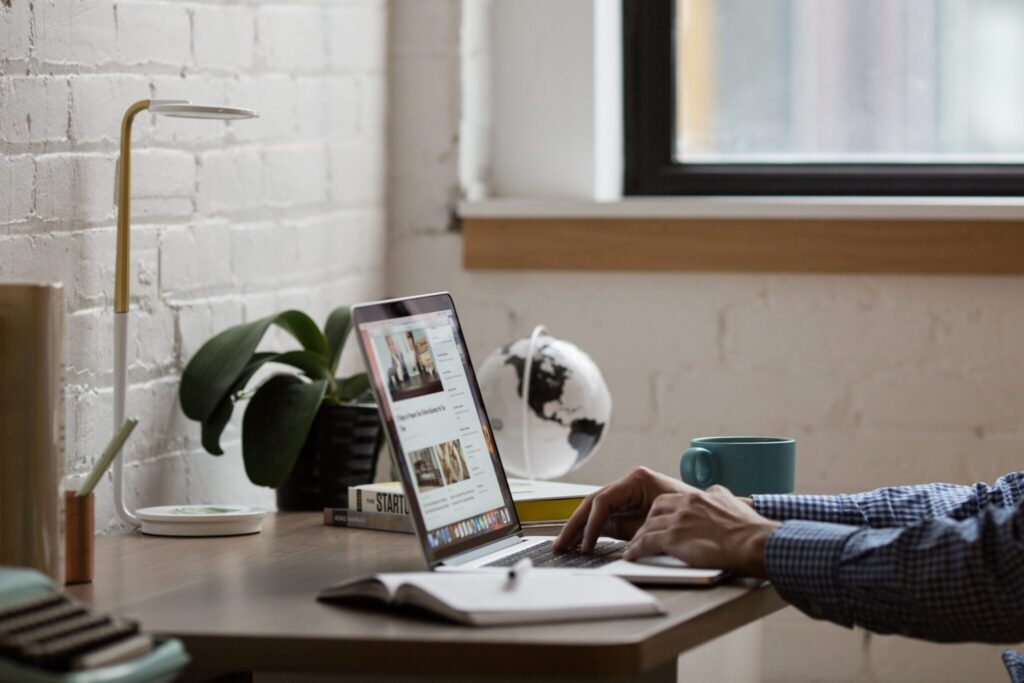 A person is working on a laptop at a wooden desk. The desk also has a potted plant, a small globe, a notepad, and a cup of coffee—perhaps they’re researching due diligence in Uruguay. Nearby are a lamp and router. A window with natural light illuminates the background.