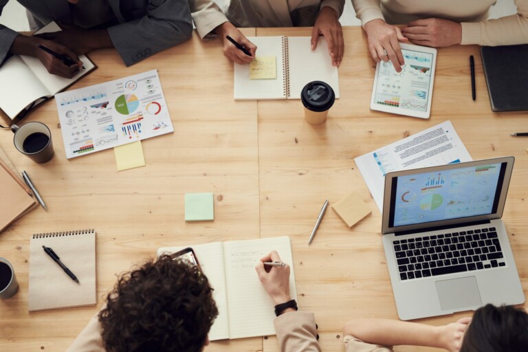 A group of people, including several caçadores de cabeças na Colômbia, sit around a wooden table with laptops, notebooks, charts, and coffee cups. They are engaged in a meeting, taking notes and discussing data and graphs displayed on papers and a laptop screen.