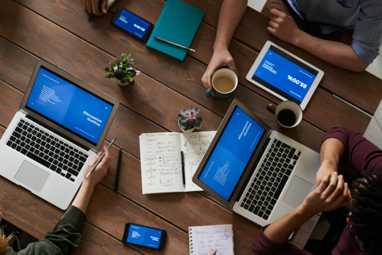 A top-down view of six people in a meeting. The table has laptops, tablets, smartphones, notebooks, and coffee cups on it. The devices display blue screens with graphs and text. One person is taking notes in a notebook while others engage with their devices.