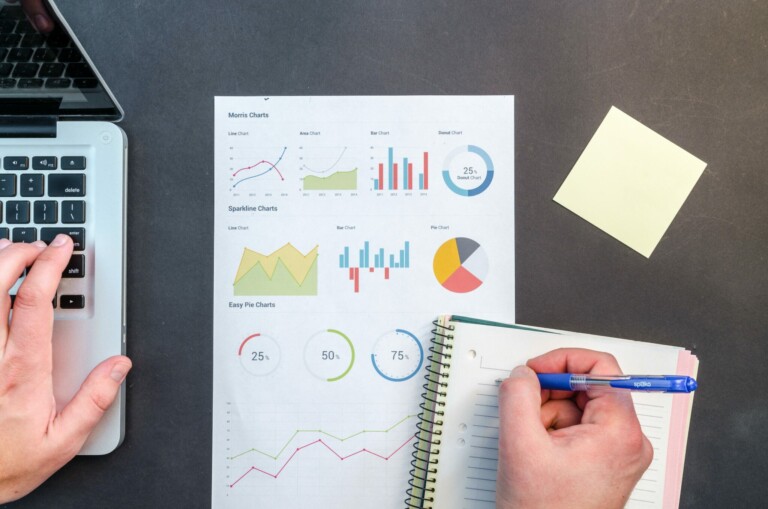 A person reviews a document with various colorful charts and graphs, including pie charts and line graphs. Their left hand uses a laptop keyboard, and their right hand writes in a spiral notebook. A yellow sticky note on the table reads "do business in El Salvador.