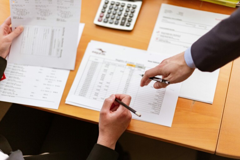 Two individuals are examining financial documents at a desk, one holding a printed sheet while the other points to a spreadsheet with a pen, likely discussing tax requirements in the Dominican Republic. A calculator and additional papers are also on the desk.